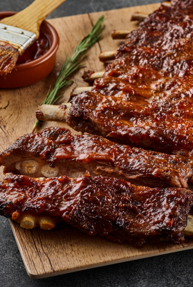 Mit Barbecue Sauce glasierte Sparerips sind auf einem Holzbrett angerichtet. Daneben liegt ein Zweig Rosmarin. Dahinter steht eine Schüssel mit Barbecue Sauce und ein Pinsel zum auftragen.