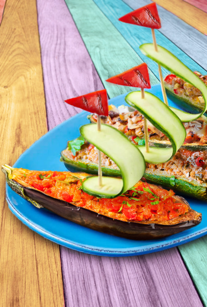 Überbackene Aubergine und überbackene Zucchini mit einem als Segel dekoriertem Spieß darin, die kindgerecht auf einem Teller angerichtet sind, welcher auf einer bunten Tischfläche steht.