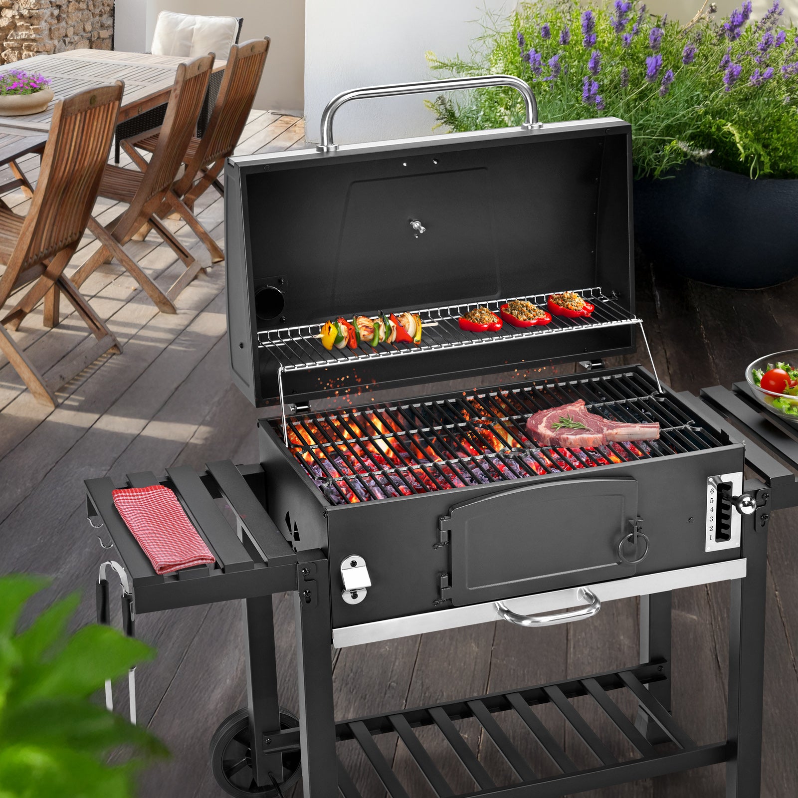 Schwarzer Grill mit offenem Deckel, auf dem Gemüse-Spieße und ein Steak gegrillt werden, steht auf einer Terrasse neben einem Holztisch mit Stühlen und blühenden Pflanzen