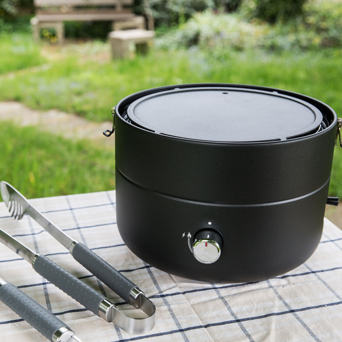 TAINO Grill-to-Go mit Grillplatte steht auf einem Tisch mit einer Grillzange daneben