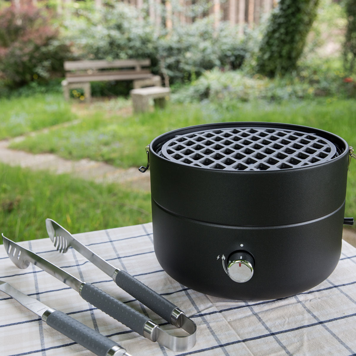 TAINO Grill-to-Go mit Grillrost steht auf einem Tisch mit einer Grillzange daneben