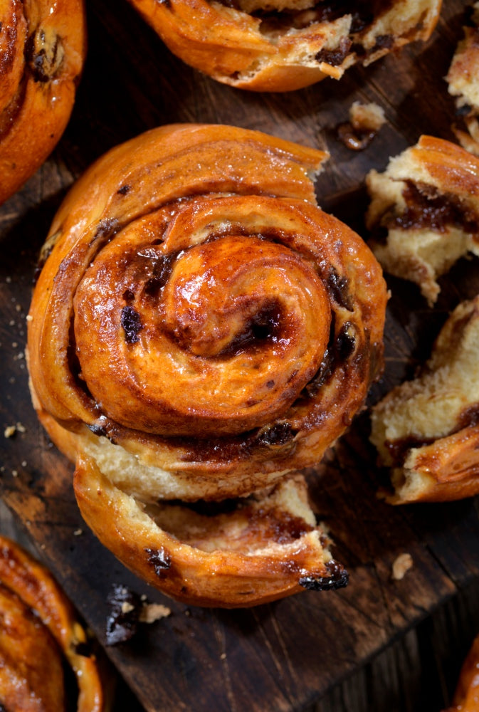 Mehrere Zimtschnecken liegen auf einem Holzbrett bereit verzehrt zu werden. Von einer Zimtschnecke ist ein Stück abgerissen, sodass man die saftige Füllung sehen kann.